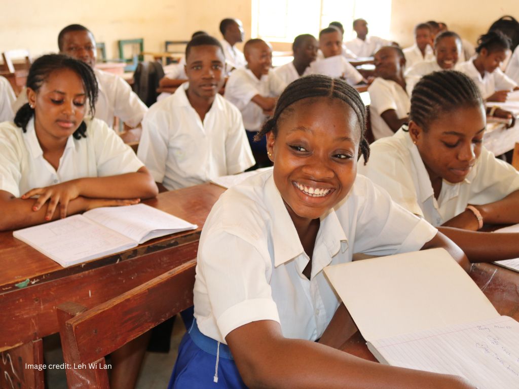 A classroom in Sierra Leone with a girl in the front wearing a white blouse, blue skirt and a big smile while she reads from a textbook