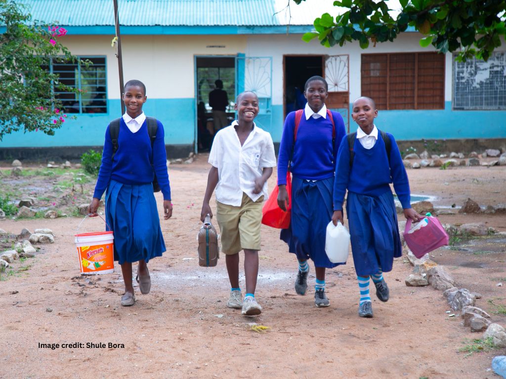 A group of smiling school children in blue school uniform and white shirts walk away from school towards the camera